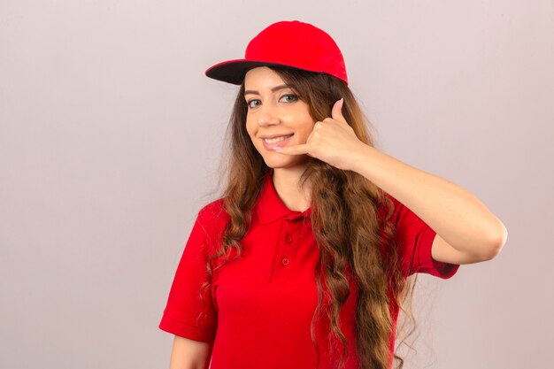 赤いポロシャツと分離の白い背景の上に自信を持って探しているジェスチャーを呼んでキャップを身に着けている若い配達の女性