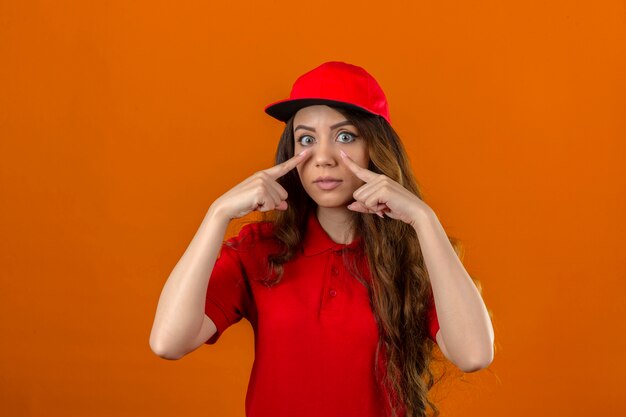 Young delivery woman wearing red polo shirt and cap making a gesture of being careful with her hands pointing at her eyes over isolated orange background