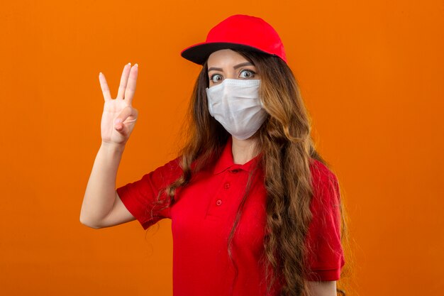 Young delivery woman wearing red polo shirt and cap in medical protective mask looking surprised doing ok sign over isolated orange background