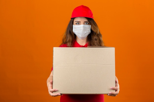 孤立したオレンジ色の背景に自信を持って見ている段ボール箱で立っている医療防護マスクで赤いポロシャツとキャップを着ている若い配達の女性