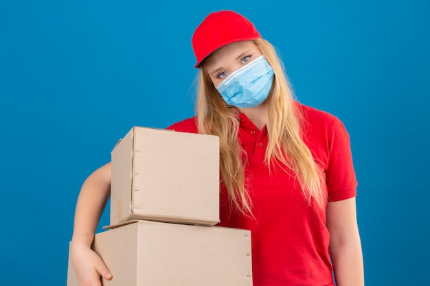 Молодая доставщица в красной рубашке поло и кепке в медицинской защитной маске стоит с картонными коробками и выглядит усталой и перегруженной на изолированном синем фоне