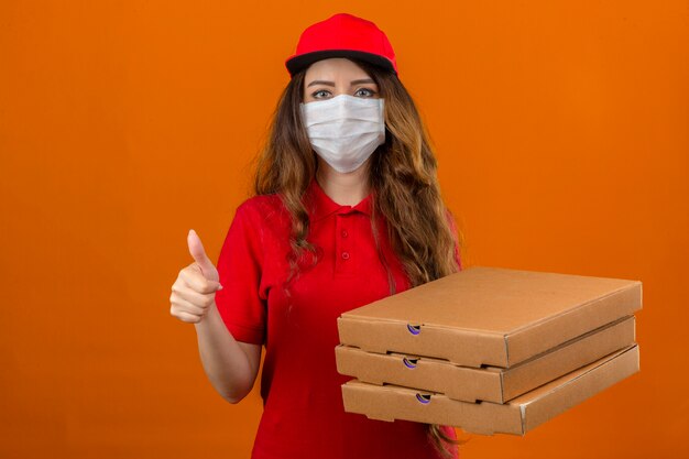 Young delivery woman wearing red polo shirt and cap in medical protective mask standing with stack of pizza boxes showing thumb up over isolated orange background