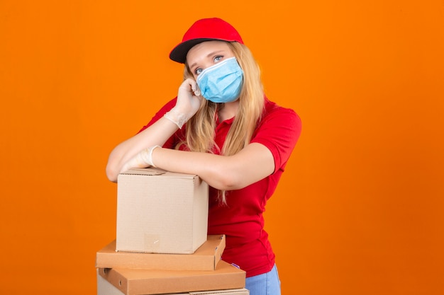 Young delivery woman wearing red polo shirt and cap in medical protective mask waiting holding hand on cheek while support it with another crossed hand with stack of cardboard boxes looking tired