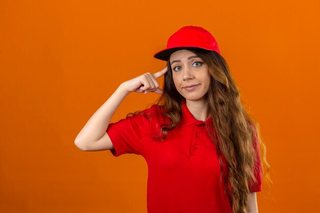 赤いポロシャツと分離のオレンジ色の背景に指の懐疑論者と寺院を指すキャップを身に着けている若い配達女性