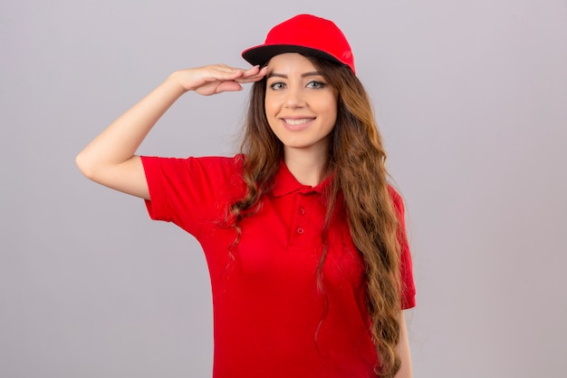 Молодая женщина-доставщик в красной рубашке поло и кепке салютует, глядя в камеру, дружелюбно улыбаясь на изолированном белом фоне