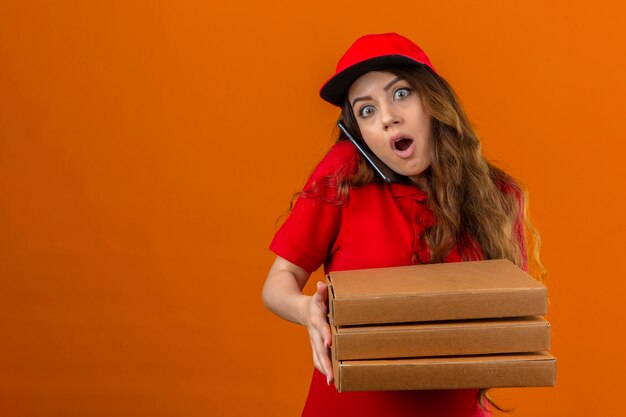 Young delivery woman wearing red polo shirt and cap shocked standing with stack of pizza boxes with wide opened eyes talking on mobile phone over isolated orange background