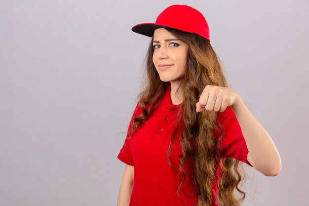 Молодая курьерская женщина в красной рубашке поло и кепке показывает кулак, дружелюбно улыбаясь на изолированном белом фоне