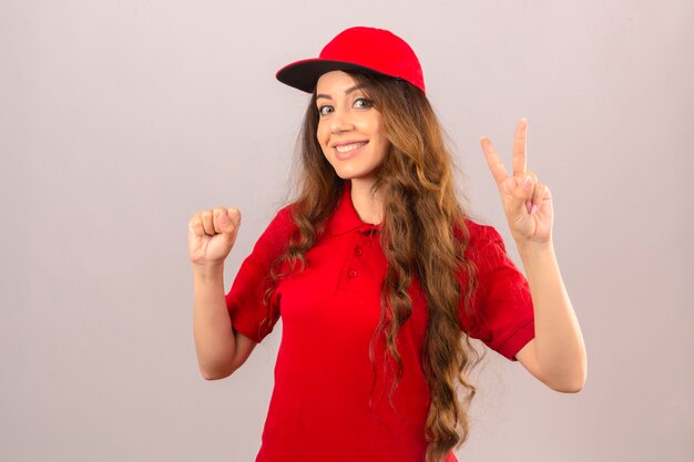 赤いポロシャツと笑みを浮かべて、孤立した白い背景の上の勝利のサインを示すキャップを着ている若い配達の女性