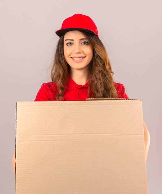 Young delivery woman wearing red polo shirt and cap standing with cardboard box smiling cheerfully over isolated white background