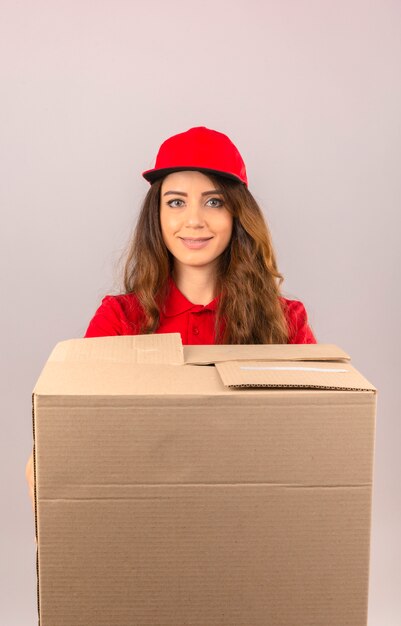 격리 된 흰색 배경 위에 유쾌 하 게 웃 고 골 판지 상자와 함께 빨간색 폴로 셔츠와 모자 서 입고 젊은 배달 여자