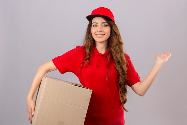 赤いポロシャツと段ボール箱に立っているキャップを着ている若い配達の女性は元気に笑みを浮かべて提示し、孤立した白い背景の上にカメラを見て手のひらで指して