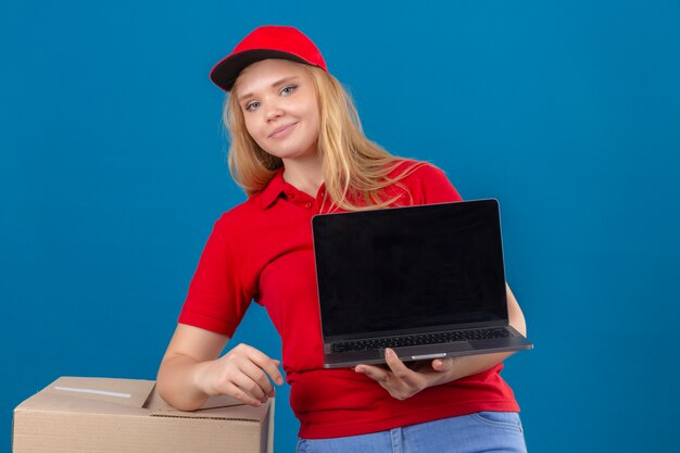 Young delivery woman wearing red polo shirt and cap standing with laptop looking confident with smile on face over isolated blue background