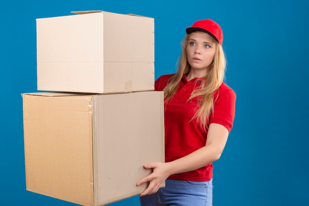 Молодая женщина-доставщик в красной рубашке поло и кепке стоит с большими картонными коробками и удивленно смотрит на изолированный синий фон