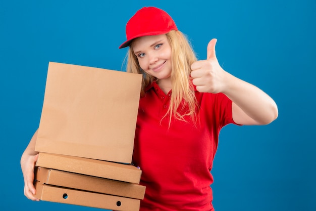 Молодая доставщица в красной рубашке поло и кепке стоит с коробками для пиццы и бумажным пакетом, показывая большой палец вверх, дружелюбно улыбаясь на изолированном синем фоне