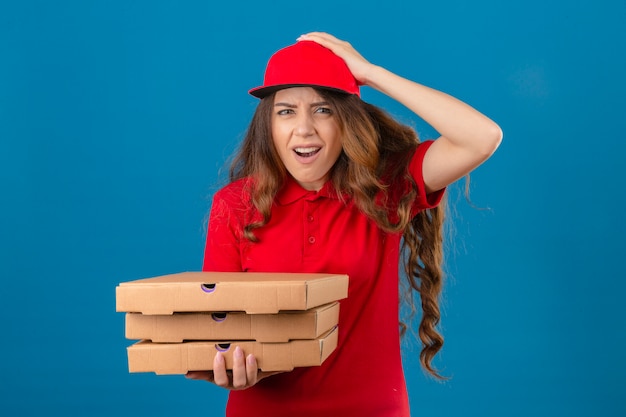 Young delivery woman wearing red polo shirt and cap standing with pizza boxes shocked with hand on head for mistake remember error forgot bad memory concept over isolated white background