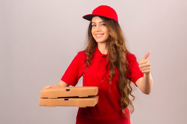 Young delivery woman wearing red polo shirt and cap standing with stack of pizza boxes smiling showing thumb up over isolated white background