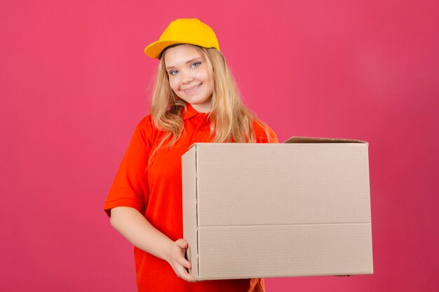 Young delivery woman wearing red polo shirt and yellow cap holding large cardboard box smiling friendly with happy face over isolated pink background