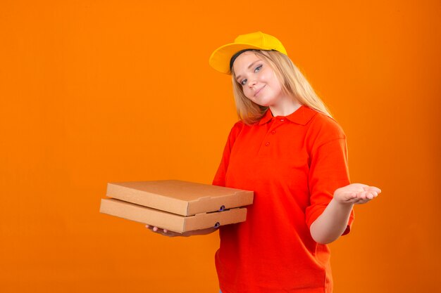 Молодая курьерская женщина в красной рубашке поло и желтой кепке держит коробки для пиццы и смущенно пожимает плечами на изолированном оранжевом фоне