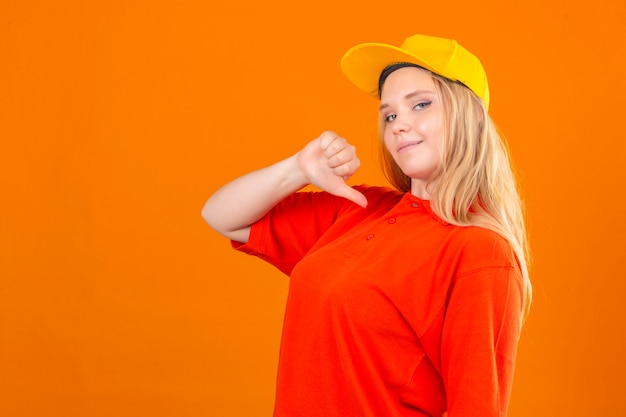 Молодая курьерская женщина в красной рубашке поло и желтой кепке уверенно смотрит на себя, улыбаясь на изолированном оранжевом фоне