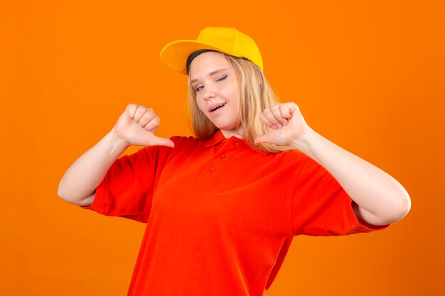 Young delivery woman wearing red polo shirt and yellow cap looking confident pointing at herself winking over isolated orange background