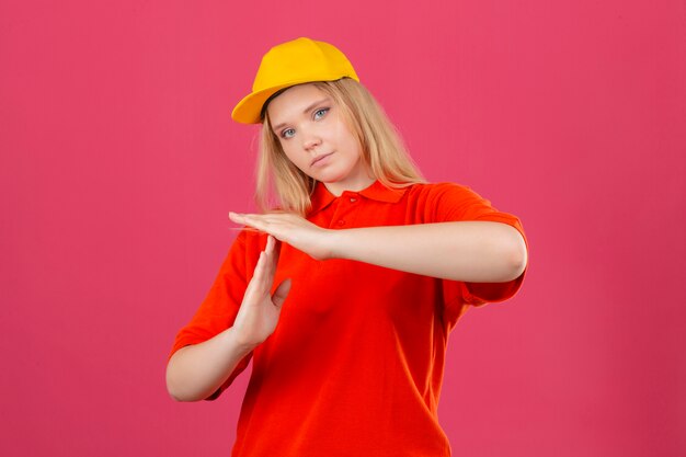 Молодая курьерская женщина в красной рубашке поло и желтой кепке выглядит перегруженной, делая жест тайм-аута руками на изолированном розовом фоне