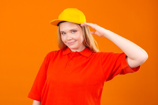 Young delivery woman wearing red polo shirt and yellow cap saluting looking confident over isolated orange background