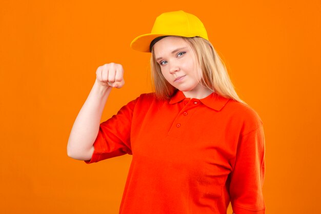 Молодая женщина-доставщик в красной рубашке поло и желтой кепке улыбается, дружелюбно стоит с поднятым кулаком победителя концепции на изолированном оранжевом фоне
