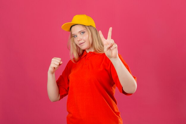赤いポロシャツと笑みを浮かべて拳を上げると分離のピンクの背景に勝利のサイン勝者の概念を示す黄色の帽子を着ている若い配達の女性
