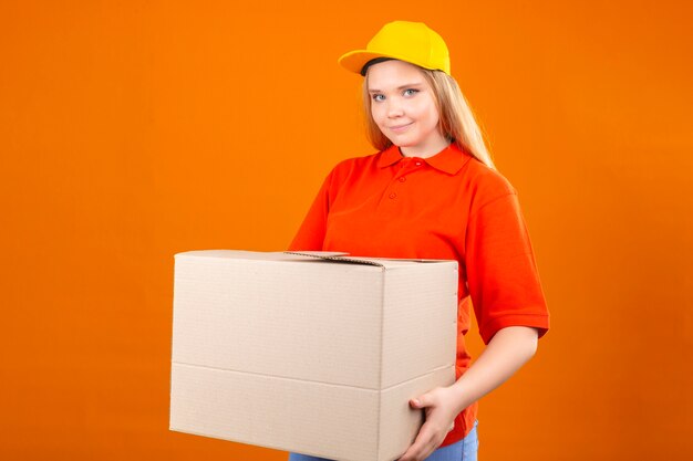 Young delivery woman wearing red polo shirt and yellow cap standing with cardboard box smiling friendly over isolated orange background