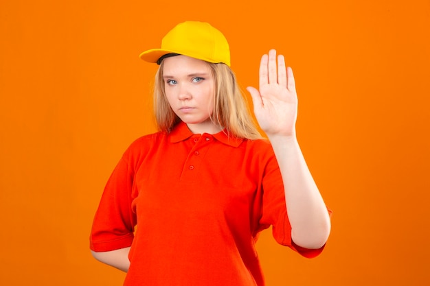 Молодая доставщица в красной рубашке поло и желтой кепке, стоящая с открытой рукой, делает знак остановки с серьезным и уверенным жестом защиты на изолированном оранжевом фоне