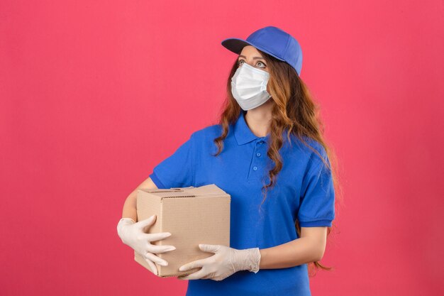 Молодая женщина-доставщик с вьющимися волосами, одетая в синюю рубашку поло и кепку в медицинской защитной маске и перчатках, глядя вверх, стоя с картонной коробкой на изолированном розовом фоне