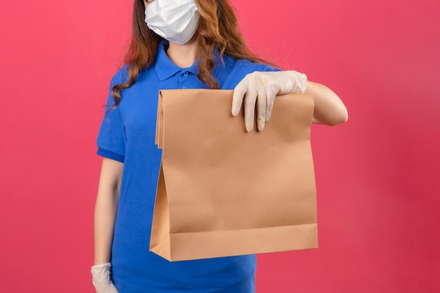 Giovane donna di consegna con capelli ricci che indossa la maglietta polo blu e cappuccio in maschera protettiva medica e guanti in piedi con il pacchetto di carta su sfondo rosa isolato