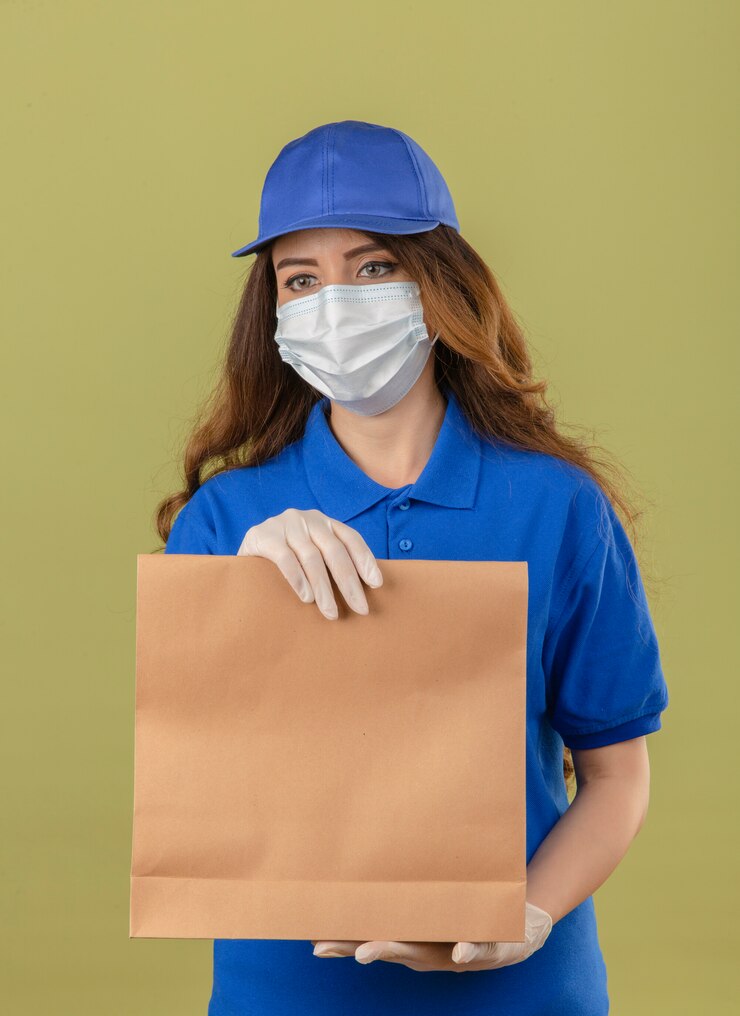 Free Photo young delivery woman with curly hair wearing blue polo shirt and cap in medical protective mask standing with paper package looking aside with serious face over isolated green background