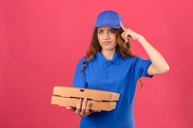 Young delivery woman with curly hair wearing blue polo shirt and cap showing a disappointment gesture with forefinger over isolated pink background