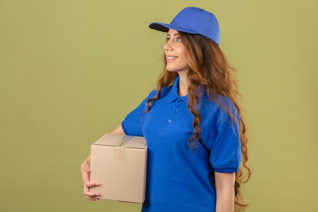 Young delivery woman with curly hair wearing blue polo shirt and cap standing sideways with cardboard box smiling over isolated green background