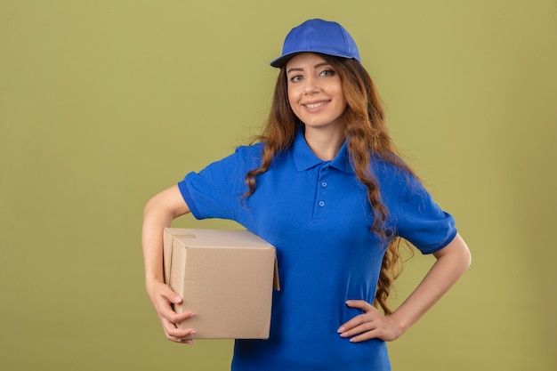 Молодая женщина-доставщик с вьющимися волосами, одетая в синюю рубашку поло и кепку, стоя с картонной коробкой, глядя в камеру, дружелюбно улыбаясь на изолированном зеленом фоне