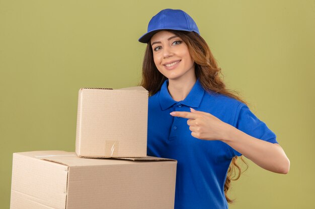 Young delivery woman with curly hair wearing blue polo shirt and cap standing with cardboard boxes pointing with finger smiling over isolated green background