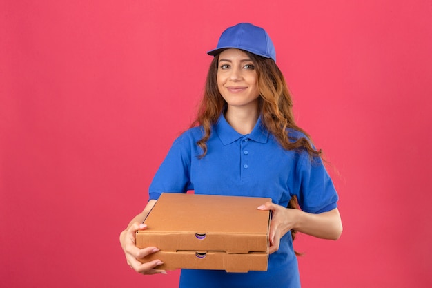 Young delivery woman with curly hair wearing blue polo shirt and cap standing with pizza boxes looking confident over isolated pink background