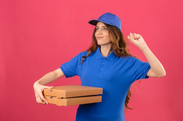 Young delivery woman with curly hair wearing blue polo shirt and cap standing with pizza boxes raising fist after a victory happy face winner concept over isolated pink background