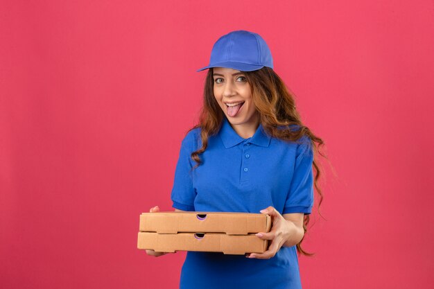 Молодая женщина-доставщик с вьющимися волосами, одетая в синюю рубашку поло и кепку, стоя с высунутым языком коробки для пиццы, развлекаясь на изолированном розовом фоне
