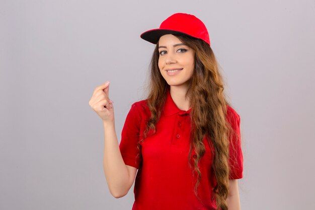 Young delivery woman with curly hair wearing red polo shirt and cap doing a money gesture smiling friendly over isolated white background