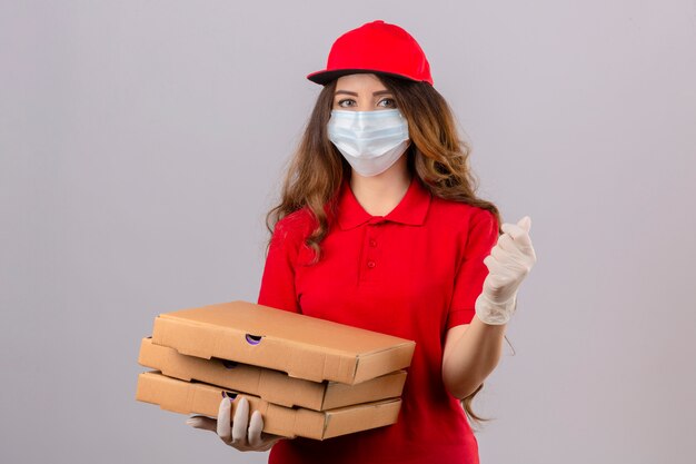 Young delivery woman with curly hair wearing red polo shirt and cap in medical protective mask and gloves standing with pizza boxes doing a money gesture smiling over isolated white background