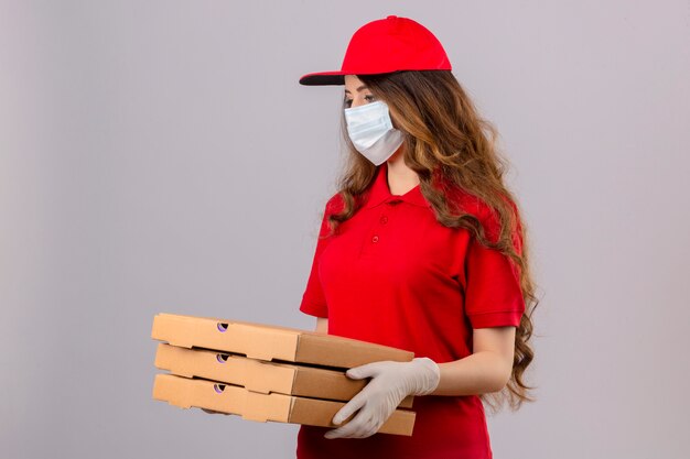 Молодая доставщица с вьющимися волосами в красной рубашке поло и кепке в медицинской защитной маске и перчатках стоит с коробками для пиццы, выглядящими очень грустными и несчастными на изолированном белом фоне