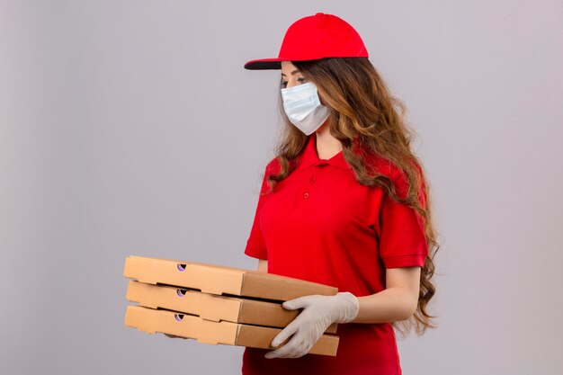 Young delivery woman with curly hair wearing red polo shirt and cap in medical protective mask and gloves standing with pizza boxes looking very sad and unhappy over isolated white background