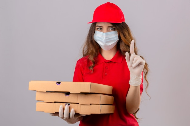 赤いポロシャツと医療防護マスクと分離の白いれたらに深刻な顔で指を上向きにしてピザの箱で立っている手袋でキャップを着ている巻き毛の若い配達の女性
