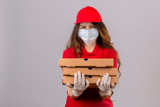 Молодая женщина-доставщик с вьющимися волосами, одетая в красную рубашку поло и кепку в медицинской защитной маске и перчатках, стоит с коробками для пиццы, скептически и нервно нахмурившись, расстроена из-за проблемы с изолой
