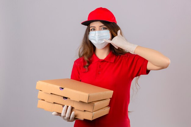 Young delivery woman with curly hair wearing red polo shirt and cap in medical protective mask and gloves with pizza boxes making call me gesture looking confident over isolated white background