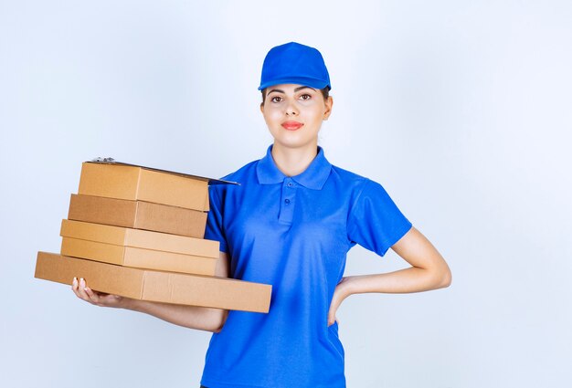 Young deliverywoman in blue uniform holding cardboard boxes on white background.