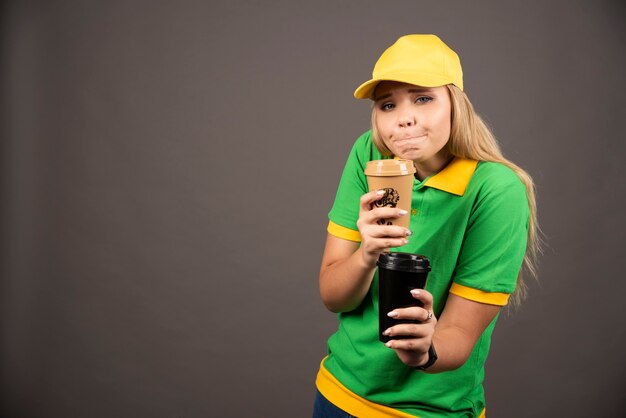 Young deliverywoman holding cups of coffee on black wall.