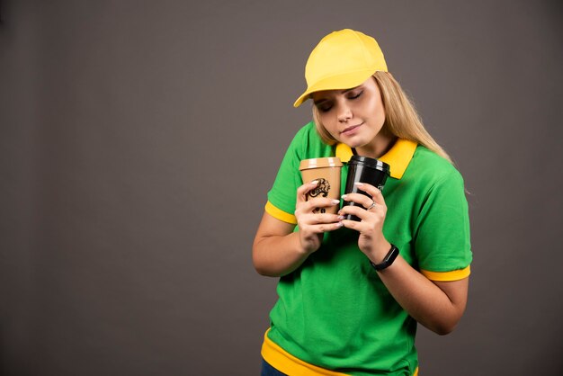 Young deliverywoman holding cups of coffee on black wall.